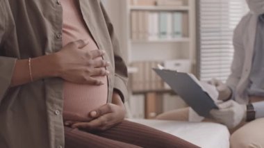 Tanımlanamayan hamile bir kadının doktor koltuğunda otururken karnını okşamasının yavaş çekimleri.
