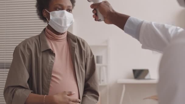 若いアフリカ系アメリカ人妊娠中の女性の傾き遅いショットは彼女の腹を撫でながら男性医師は診療所で任命を持っている非接触赤外線温度計で彼女の温度を測定 — ストック動画