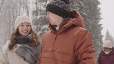Karlı bir günde genç beyaz bir çiftin arkadaşlarıyla birlikte güzel bir kış ormanı boyunca yürüdüğü orta yavaş bir çekim.