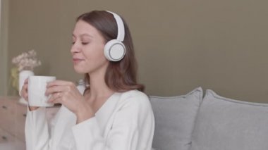 Kulaklıkla müzik dinleyen, çay içen, akıllı telefondan kayan, rahat bir evde yatağında oturan genç ve rahat hamile bir kadının orta yavaş çekimi.