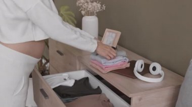 Müstakbel annenin karnını şefkatle okşayarak minik bebek kıyafetlerini çekmeceye koyduğu yavaş çekim.