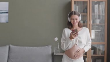 Elinde akıllı telefonuyla kulaklıkla müzik eşliğinde dans eden ve karnını okşayan mutlu hamile kadının orta yavaş çekimi.