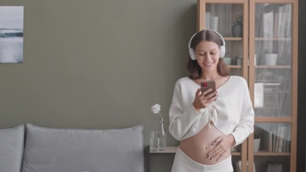 拿着智能手机的快乐的年轻孕妇手持耳机在音乐中跳舞 抚摸着她的肚子 拍了一张中等速度的照片 — 图库视频影像