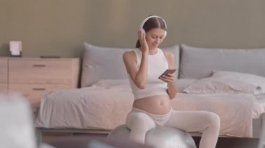 Evde spor salonunda oturmuş kulaklıkla müzik dinleyen gülümseyen hamile bir kadının yavaş çekimi.