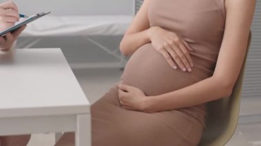 Modern doktor muayenehanesinde randevu sırasında doktorla konuşurken dar bej elbiseli, hamile bir kadının göbeğini tutarken çekilen yavaş çekim.