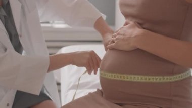 Dar bej elbiseli, tanınmayan hamile bir kadının göbek ölçüsünü alan doktorun yavaş yavaş yakın çekimi.