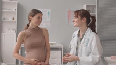 Genç Kafkas hamile kadın ve kadın doğum uzmanının doktor muayenehanesindeki randevularında konuştuğu ve sonra birlikte kameraya gülümsediği orta yavaş bir çekim.