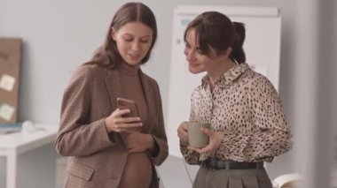 İş yerindeki kadın meslektaşına bebek ultrasonu resimlerini gösterirken karnını okşayan mutlu, genç, beyaz bir kadının orta yavaş çekimi.