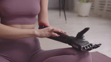 Evde yoga yaparken yüksek teknolojili protez kolunu kullanan, mor aktif giysili, tanınmayan genç bir kadının yavaş çekimleri.