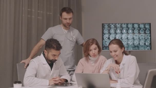 一组年轻的医生在会议室里一边看着笔记本电脑一边聊天 背景上还展示着脑部X光照片 — 图库视频影像