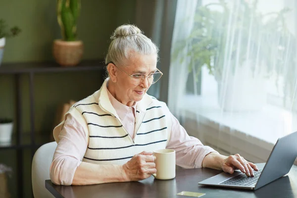 Senior mujer en ropa de casualwear teniendo té y mirando a la pantalla del ordenador portátil — Foto de Stock
