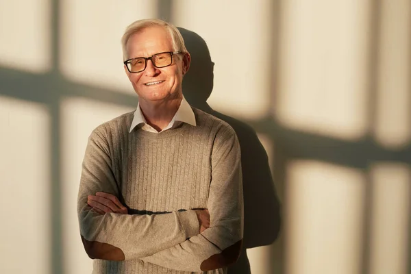 Feliz hombre mayor con el pelo gris cruzando los brazos por el pecho mientras está de pie por la pared —  Fotos de Stock