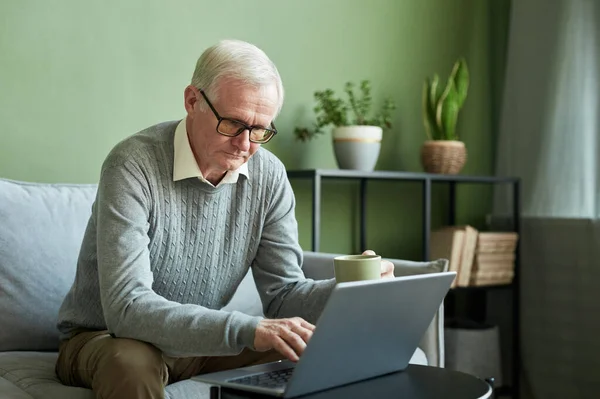 Серйозний старший чоловік у повсякденному одязі сидить на дивані перед ноутбуком — стокове фото