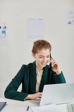 Genç neşeli iş kadını ya da sekreteri iş yerinde cep telefonuyla konuşuyor.