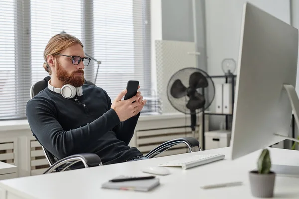 Joven hombre de negocios barbudo desplazándose en el teléfono inteligente mientras está sentado en el lugar de trabajo — Foto de Stock