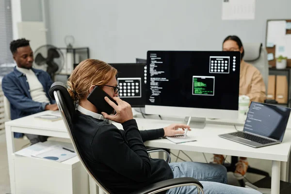 Ung upptagen programmerare talar på mobiltelefon medan du sitter framför datorn — Stockfoto