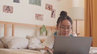 Gözlüklü Asyalı genç kızın, odasında dizüstü bilgisayarla arkadaşlarıyla mesajlaşırken gülümsediği orta boy bir fotoğraf.