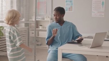 Klinikteki tıbbi randevusunda 7 yaşında beyaz bir çocuğun orta yavaş çekimi. Mavi önlüklü genç Afro-Amerikalı erkek doktor onu yumruk tokuşturarak selamlıyor