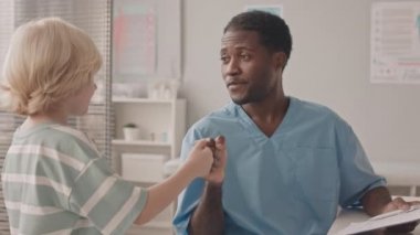 Kafkasyalı küçük bir çocuğun doktor muayenehanesine gelişinin orta yavaş çekimi. Afro-Amerikalı genç çocuk doktoru onu yumruk tokuşturarak selamlıyor.