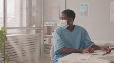 Orta yavaş çekimde maskeli genç Afro-Amerikalı doktor ve Kafkasyalı küçük çocuğun modern klinikte randevusu var, birbirlerini yumrukla selamlıyorlar.