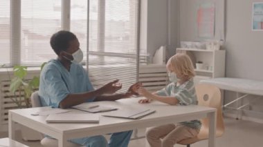 Elinde küçük bir şişe aşı tutarak aşının faydalarından bahseden genç Afrikalı-Amerikalı erkek doktoru ya da hemşireyi dinleyen 7 yaşındaki beyaz çocuğun orta yavaş çekimi.