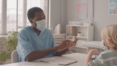Maskeli genç Afro-Amerikan doktor ve beyaz çocuğun randevusu sırasında aşılanmaktan bahsederken orta yavaş çekimleri.