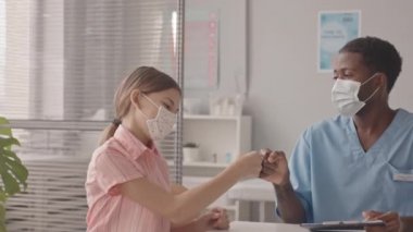 Yüz maskesi takmış 13 yaşındaki beyaz bir kızın doktorla randevusu varken, Afro-Amerikalı çocuk doktorunu yumruğuyla selamlarken, aşıya hazırlanırken orta yavaş bir çekim.