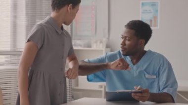 10 yaşındaki Afro-Amerikan bir kızın ofisinde doktorunu ziyaret ederken, birbirini dirsek tokuşturarak selamlarken orta yavaş bir çekim.
