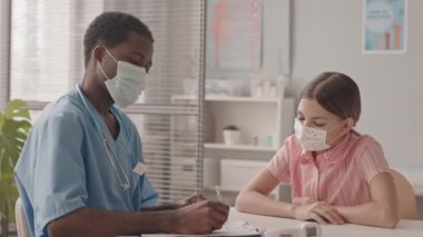 Orta yavaş çekimde, doktor ofisinde oturan beyaz yüzlü küçük bir kızın genç Afro-Amerikalı çocuk doktoruyla randevusu var.