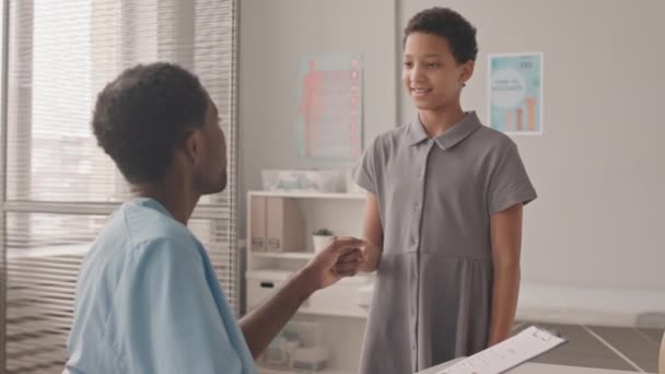 中慢镜头10岁的非洲裔美国女孩去诊所看医生 身着蓝色衣服的年轻的非洲裔美国男医生用拳头向她的友人致意 — 图库视频影像