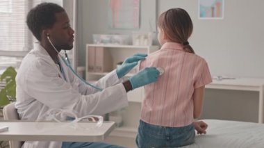 Orta yavaş çekimde, genç Afro-Amerikan erkek doktor steteskopla beyaz kız öğrenciyi muayene ediyor.