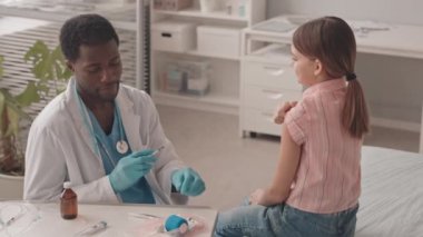 Orta yavaş çekimde, eldivenli genç Afro-Amerikan doktor beyaz kız öğrenciye tıbbi muayene koltuğunda otururken Covid-19 aşısı enjekte ediyor.