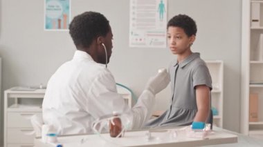Modern doktorların muayenehanesinde kanepede oturan 10 yaşındaki Afro-Amerikan bir kızın steteskopla pediatristi tarafından muayene edilirken orta boy fotoğrafı.