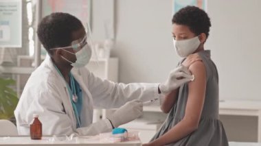 Orta boy Afro-Amerikan doktor koruyucu yüz kalkanı ve maskesiyle Afro-Amerikan okul kızına Covid-19 aşısı yaparken.