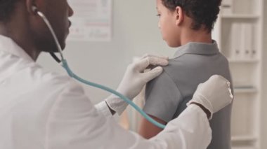 Steteskoplu Afrika kökenli Amerikalı erkek çocuk doktorunun 10 yaşındaki Afro-Amerikalı bir kızı muayene edip kalp atışlarını dinlediği orta boy yakın çekim.