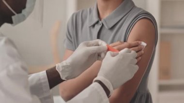 Tanımlanamayan küçük bir kızın iğne yaptıktan sonra omzuna yara bandı yapıştıran erkek doktor tarafından Covid-19 'a karşı aşılanmasının yavaş çekimleri.