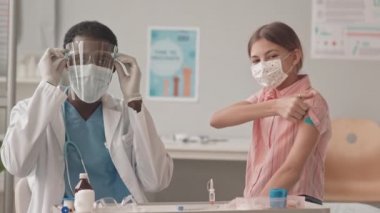 Covid-19 aşısından sonra kameraya bakan maskeli Afro-Amerikan doktor ve Kafkas öğrenci portresi. Omzunda yara bandı olan ve gülümseyen bir kız.