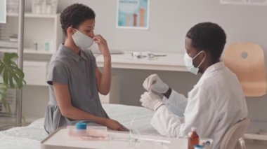 Orta yavaş çekimde, yüz maskesi takmış Afro-Amerikan bir doktorun ofisinde kanepede oturan 10 yaşındaki Afrikalı-Amerikalı bir kıza oral aşıyı verirken görülüyor.