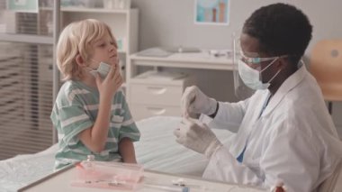 Kafkasyalı küçük bir çocuğun, genç Afro-Amerikalı erkek doktor tarafından yüz kalkanı ve maskesi takılarak doktor muayenehanesinde oral aşı olurken orta yavaş çekimleri.