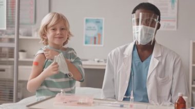 Aşı sonrası omzunda yara bandı olan beyaz bir çocuğun orta yavaş portresi. Afro-Amerikalı bir erkek doktor kameraya bakıyor.