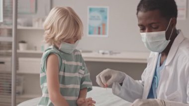 Afro-Amerikan doktorların aşı yaptıktan sonra cesur beyaz çocuğun omzuna renkli yara bandı yapıştırdığı orta yavaş bir çekim.