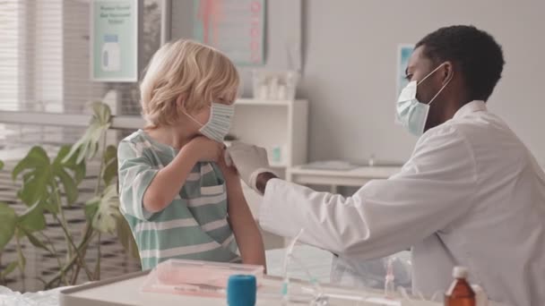 中程度の遅いショットの7歳の白人少年取得予防接種によってアフリカ系アメリカ人若い男性医師で現代クリニック — ストック動画