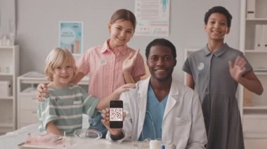 Aşılandıktan sonra doktorun ofisine el sallayan üç gülümseyen çoklu etnik çocuğun orta yavaş portresi. Afro-Amerikalı doktorları akıllı telefondan QR kodu gösteriyor.