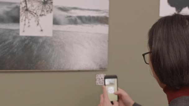 眼鏡中の長髪の青年がスマホを使ってQr コードの黒と白の抽象写真を壁に掛ける現代美術ギャラリー — ストック動画