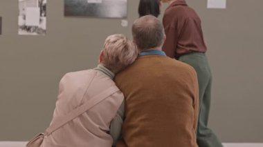 Kafası kocasının omuzunda, modern sanat galerisindeki bankta oturmuş fotoğraf sergisinin keyfini çıkaran olgun bir kadının dikiz aynasındaki yavaşlığı. Müzedeki ziyaretçilerden biri tekerlekli sandalye kullanıyor.