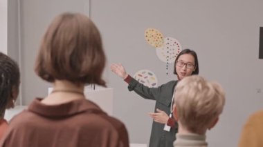Genç Asyalı erkek galericinin müzedeki sergi sırasında ziyaretçiler izlerken ve dinlerken kavramsal modern sanat objelerinden bahsederken orta yavaş çekimleri.
