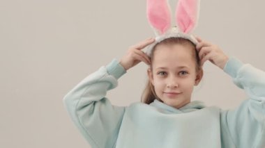 Kafasına tavşan kulağı takan ve arka planda duran kameraya gülümseyen küçük güzel bir kızın orta yavaş portresi.