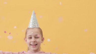 Gümüş renkli parti şapkalı, konfeti fırlatan ve parlak turuncu stüdyo arka planında duran kameraya bakan beyaz kızın yavaş yavaş portresi.