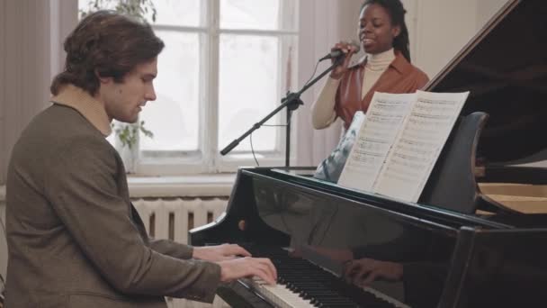 屋内で音楽を演奏する若い異人種間のカップルの中低速 白人男性はピアノを弾き アフリカ系アメリカ人女性は音楽学校でマイクを歌っている — ストック動画