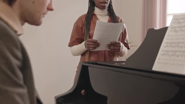中等速度的非洲裔美国年轻女歌手 手拿着歌词演唱 而年轻的白人男子弹奏着宏伟的钢琴 在音乐会前排练 — 图库视频影像